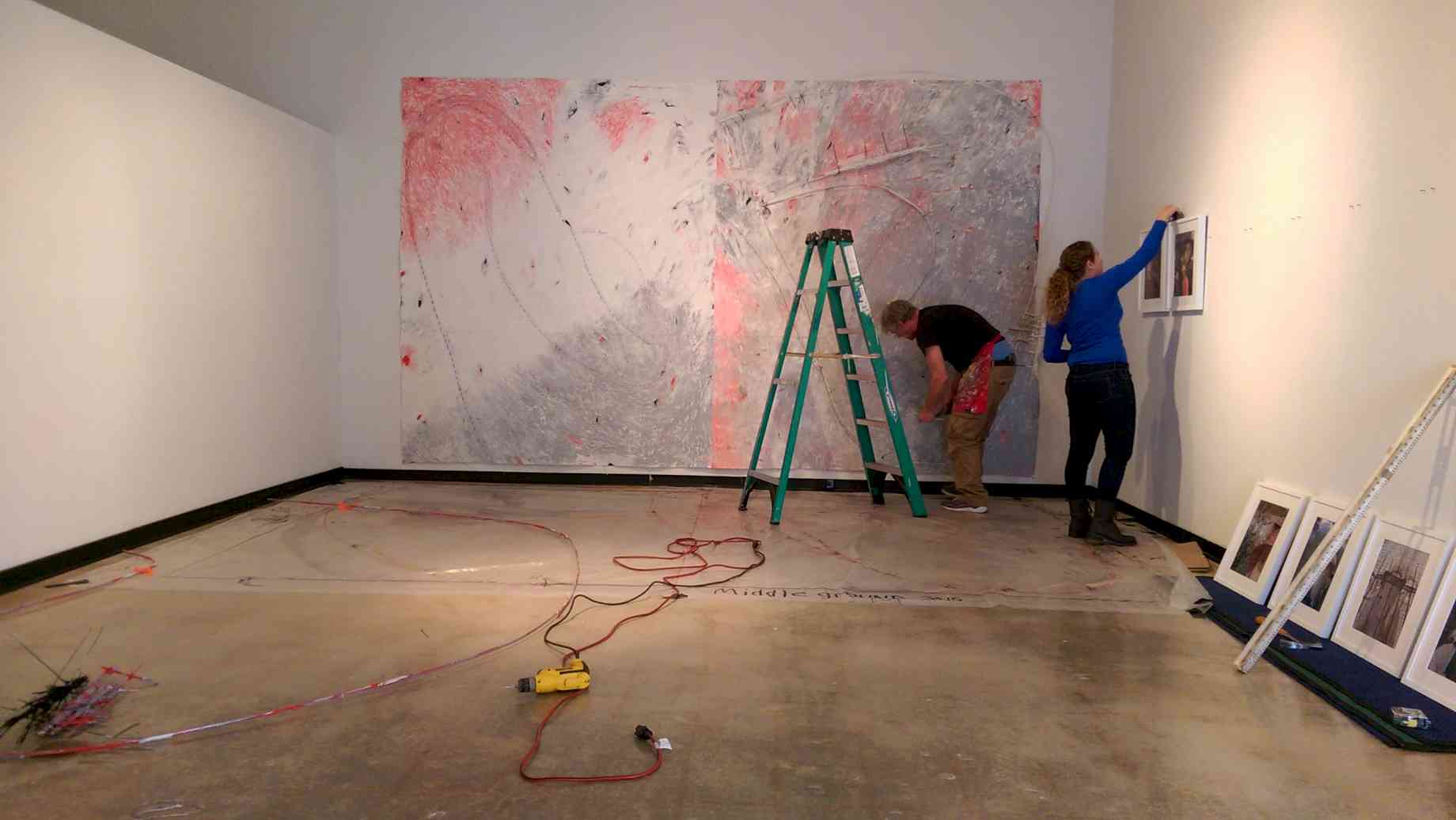 Alumni Kaitlin Hof-Mahoney installs an exhibit at the University Gallery. [Alt text: A gallery mid-installation. A woman measures two pieces of art on a wall, and a man works on a wall-sized mural.]