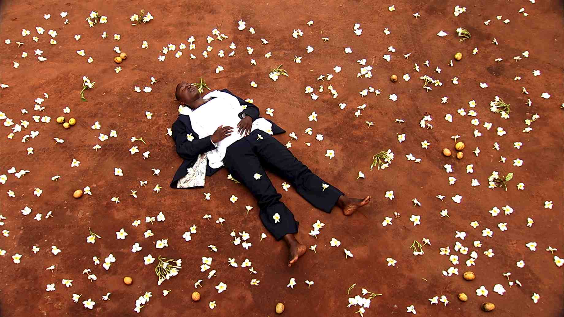 Nora Chipaumire, as her father in the film NORA, lies in stillness as flowers fall around her.