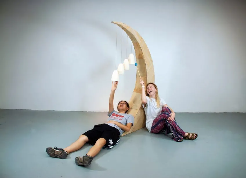 Two people interact with a musical wood sculpture of a crescent moon.
