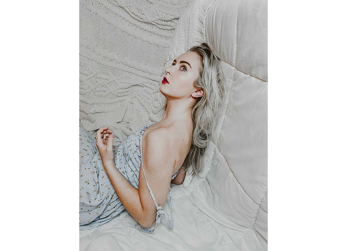 A woman wearing a sun dress and bright red lipstick laying on her side on light gray bed linens