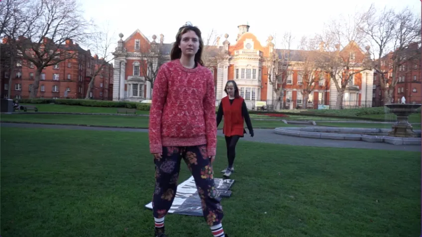 Video still. Two people standing in an urban park setting.