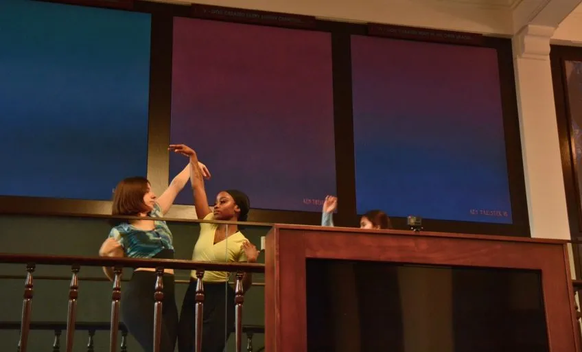 Two dancers performing on a balcony, facing each other, each with one arm reaching toward the other.