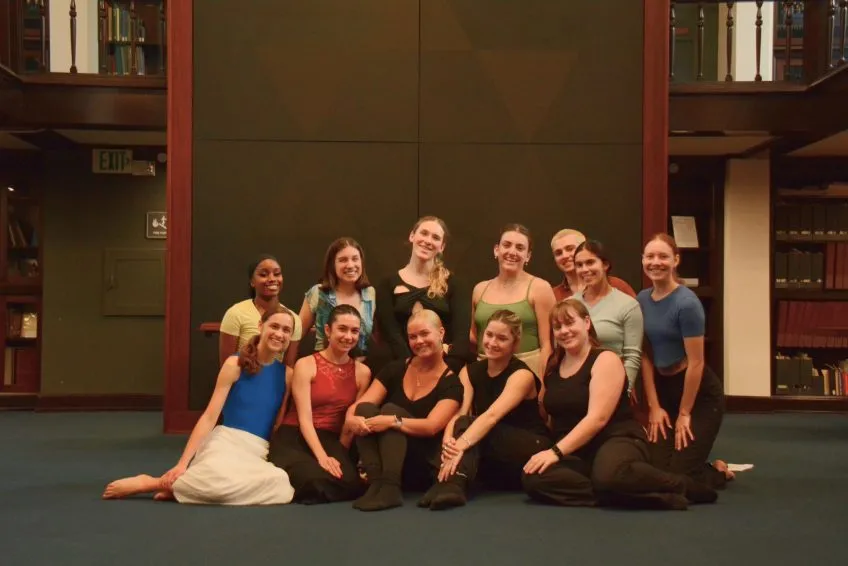 A group of twelve people in dance attire seated in a library.