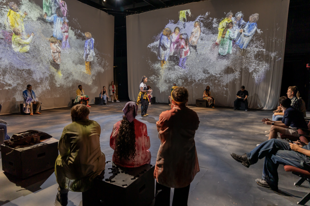 A group of people in an immersive theatre setting with the audience on stage and projections on the surrounding walls.
