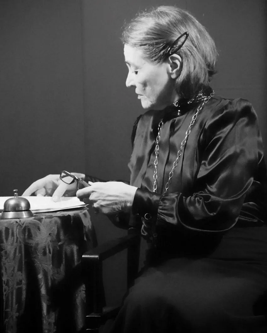 Margaret Bachus in period costuming, seated on a theatre stage looking at a book. Black and white photo.