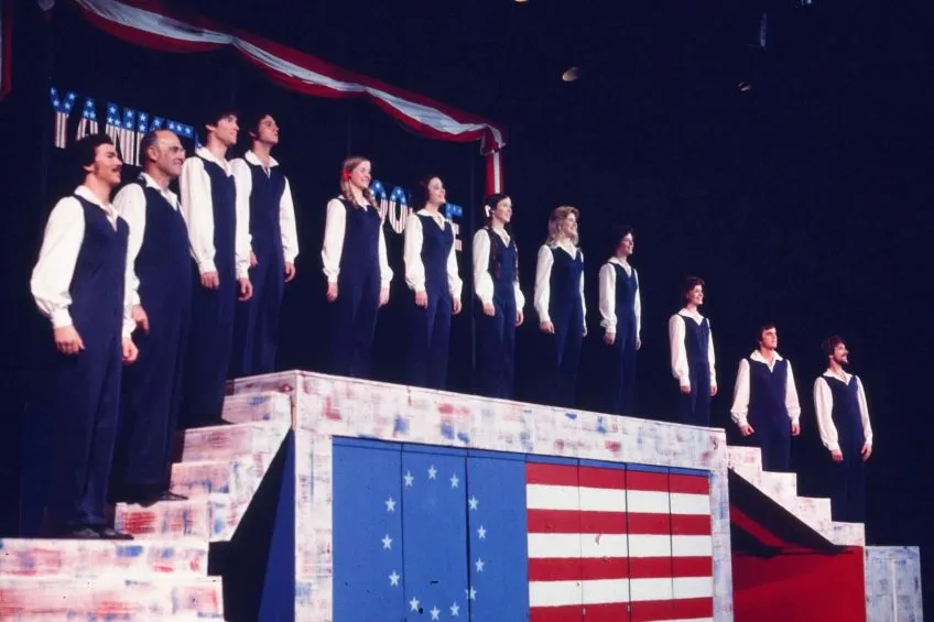 A group of actors on a stage with a Betsy Ross Revolutionary War era U.S.A. flag.