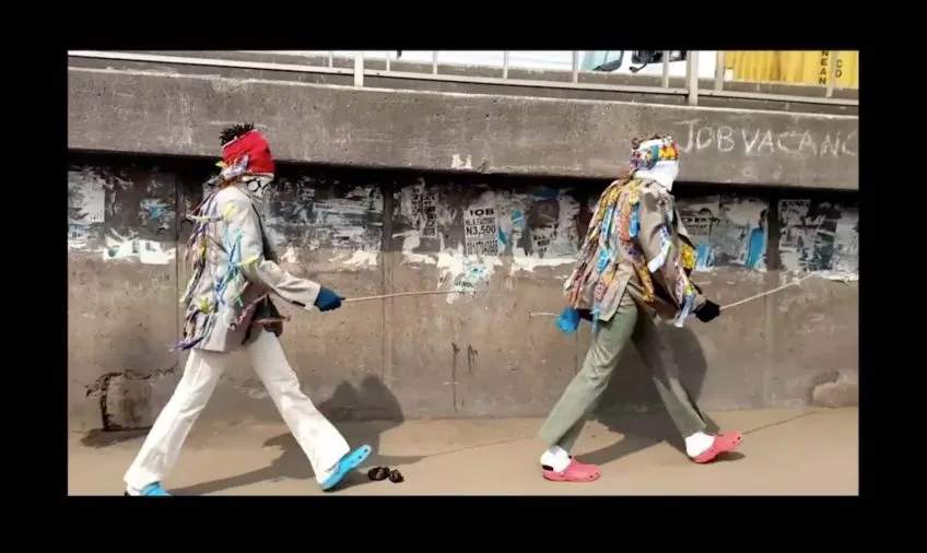 Screenshot from danceGATHERING 2020 where two people walk near a concrete wall with hands and face fully covered.