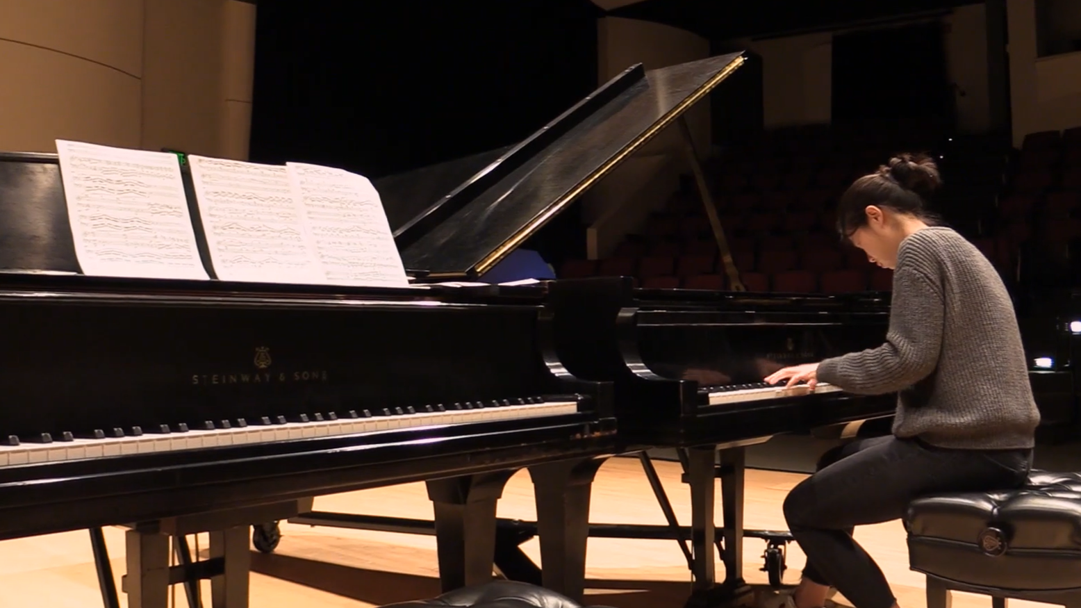 A woman plays piano on a stage. Video still.