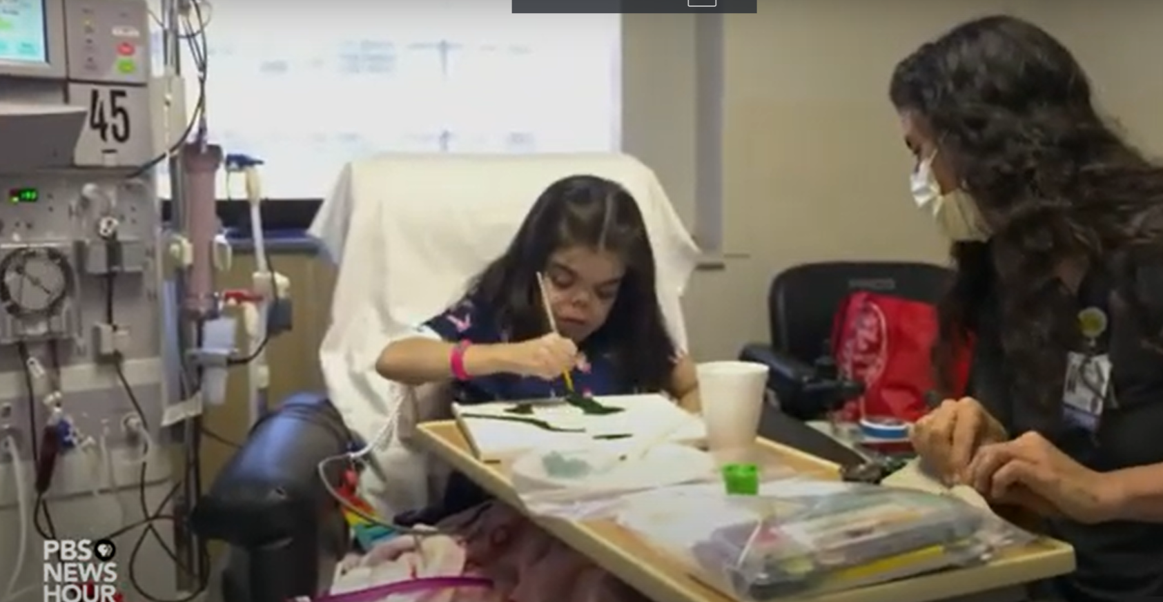 A patient in a hospital setting is painting a watercolor picture with an arts in health practitioner seated at their bedside.