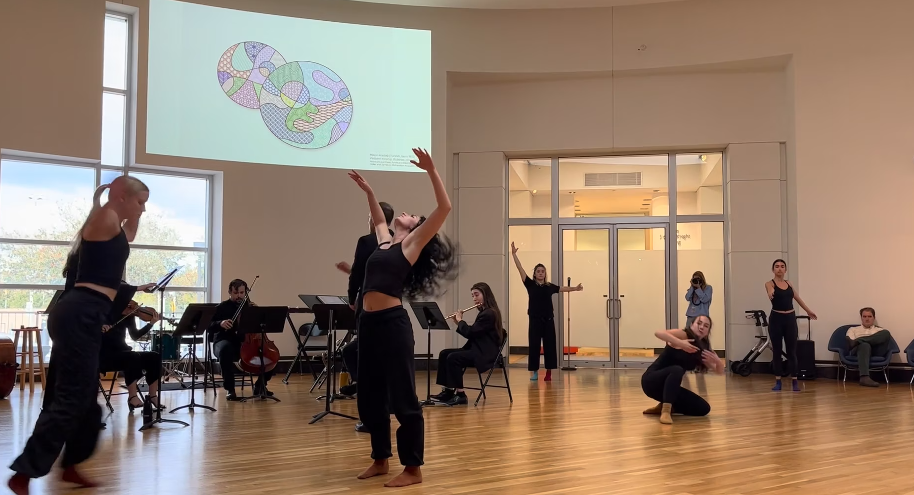 A group of people perform contemporary dance while a small music ensemble plays inside an art gallery setting
