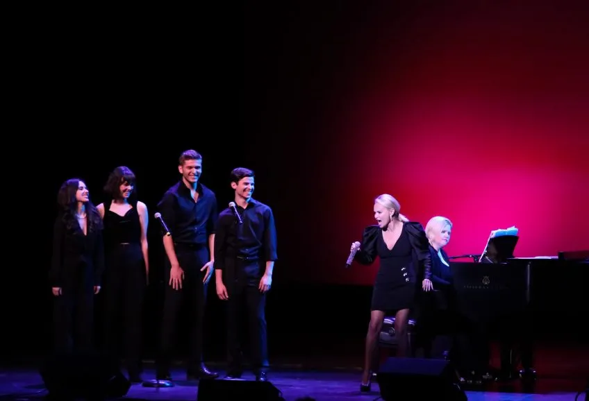 A theatre stage. Kristen Chenoweth sings while a group of students watches.