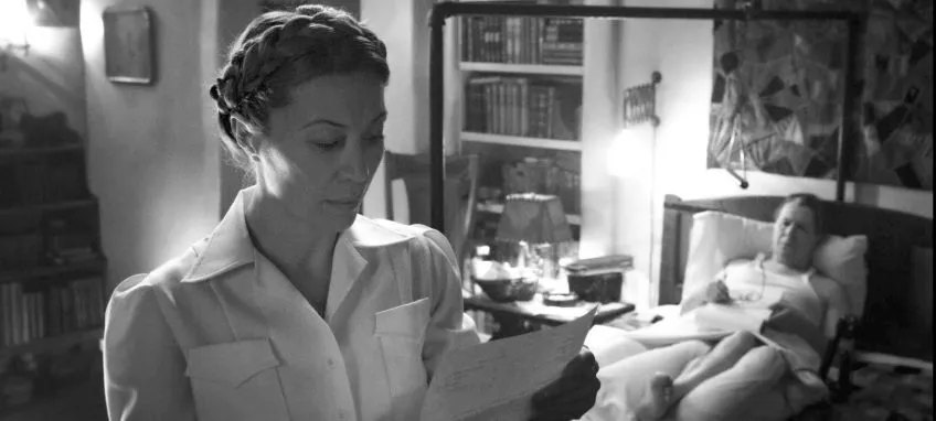 Monika Gossmann, standing and reading a paper while another character lays on a bed behind her