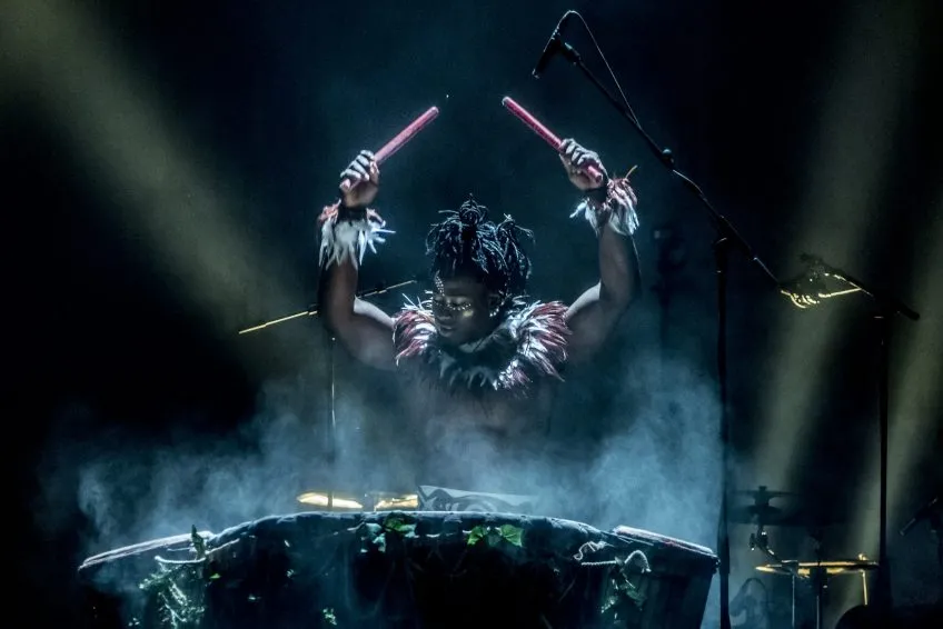 A person playing drums, wearing West African makeup, accessories, and costuming