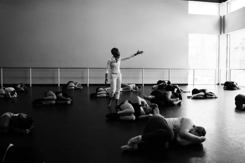 Black and white photo. At center: a dance teacher stands with arm outstretched. Dance students lie in fetal position on the floor.