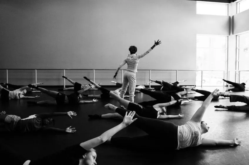 Black and white photo. At center: a dance teacher stands with arm outstretched. Dance students lie in fetal position on the floor.