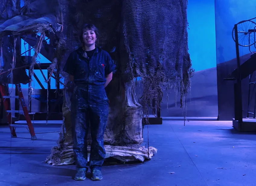 A person wearing coveralls stands on a theatre set with a prop resembling a makeshift fort