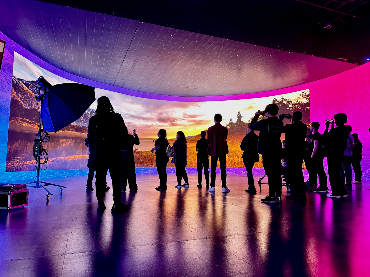 Digital Worlds students stand on a virtual production stage in front of a high-def, curved panoramic screen displaying a mountain lake sunrise