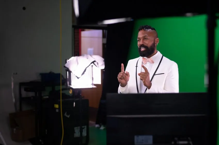 A person in a white tuxedo and pink bowtie stands in front of a green screen.