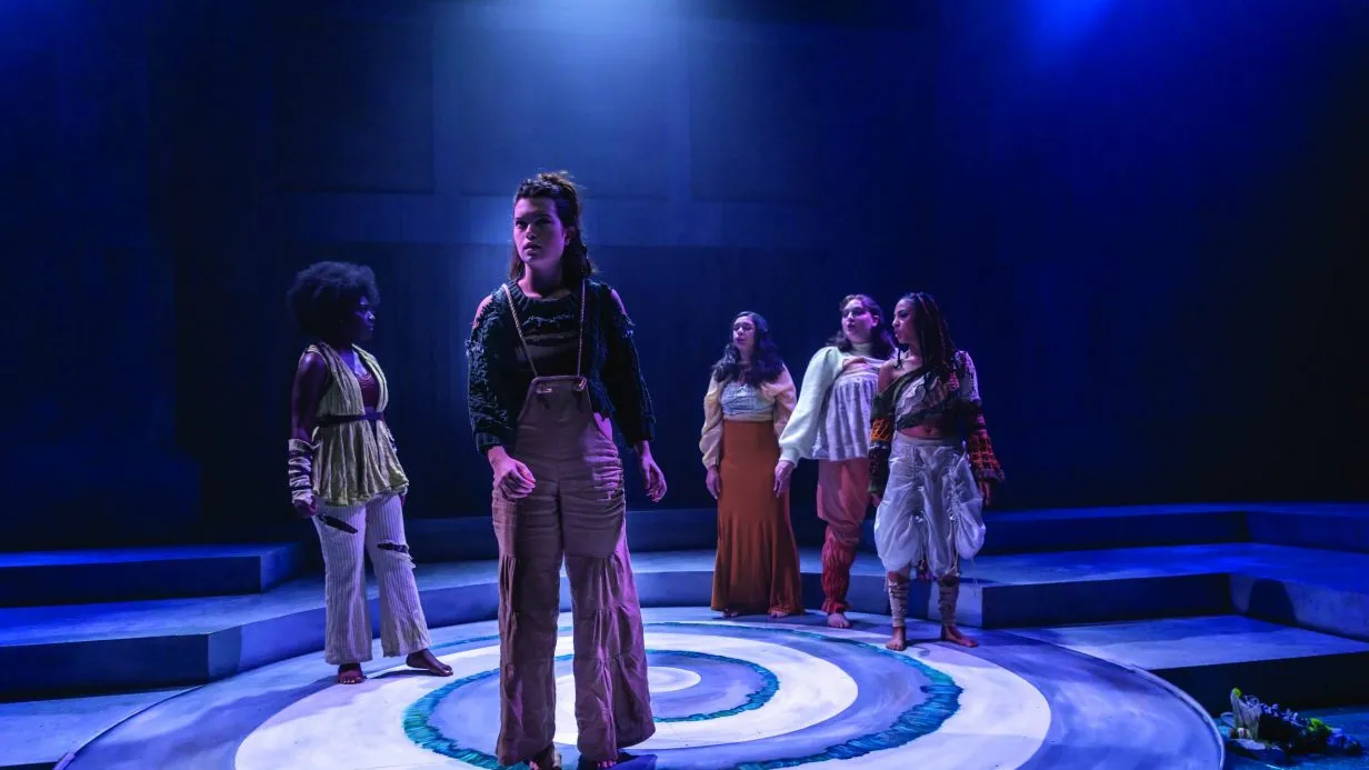 A group of actors perform under a blue spotlight on a black box theatre stage.