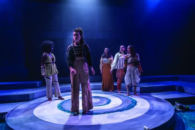 A group of actors perform under a blue spotlight on a black box theatre stage.