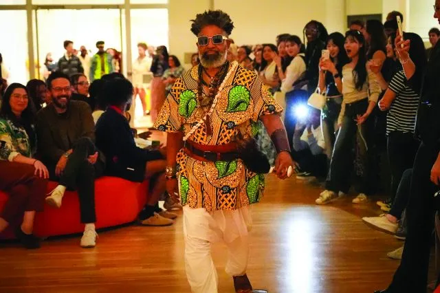 Afrofuturism fashion show. Onlookers watch a man wearing a batik fabric tunic with yellow, green and royal blue patterns, sunglasses and beaded, shell, and leather accessories do a runway walk.