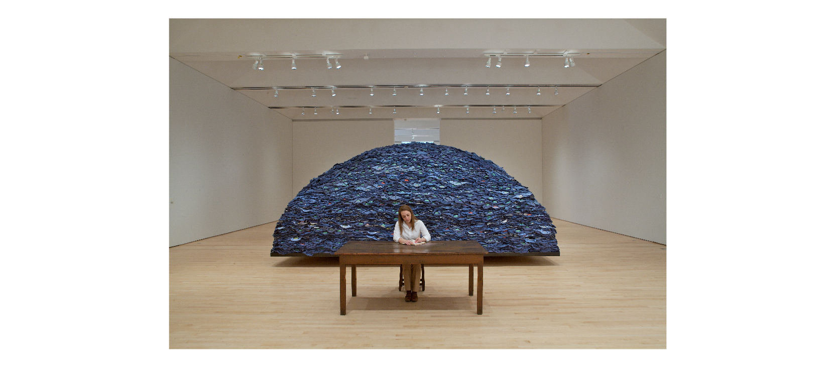 A woman seated at a table in a gallery installation -performance. Behind her a hill-shaped large pile of hundreds of blue jeans.
