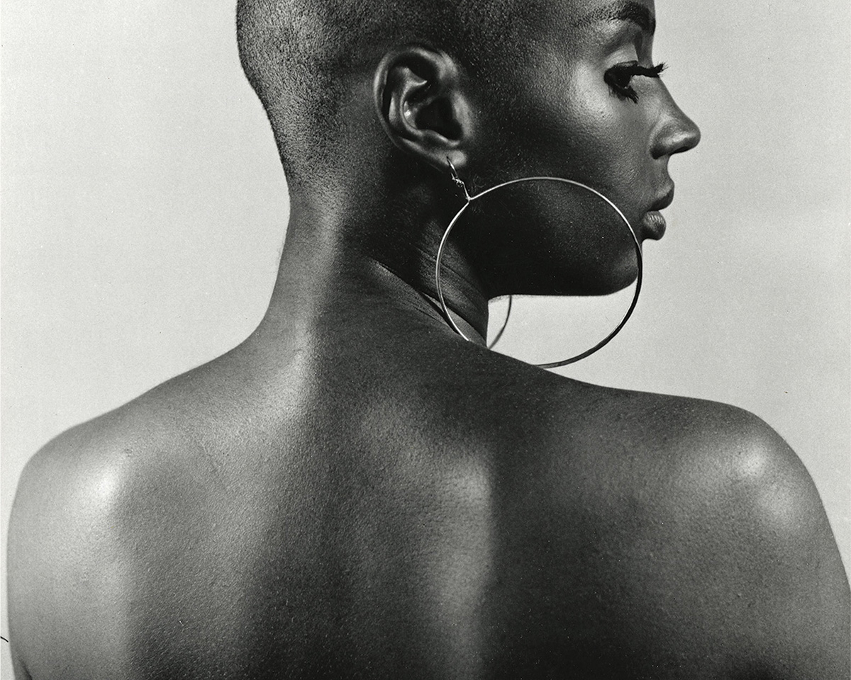 A Black woman with shaved head, hoop earrings, pictured from her bare shoulders up from behind, turning her head in profile.