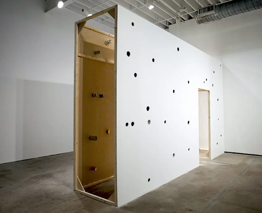 Image description: Art installation of a narrow wooden structure. Cans are embedded in walls and attached by strings criss-crossing the interior.