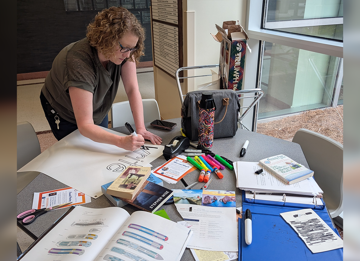 A woman writing on a posterboard on a table covered with art supplies and books.