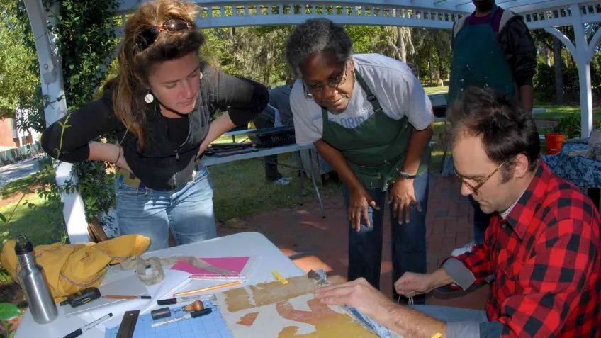 Two people standing look on as a third person sitting works on a craft project at a table outside.