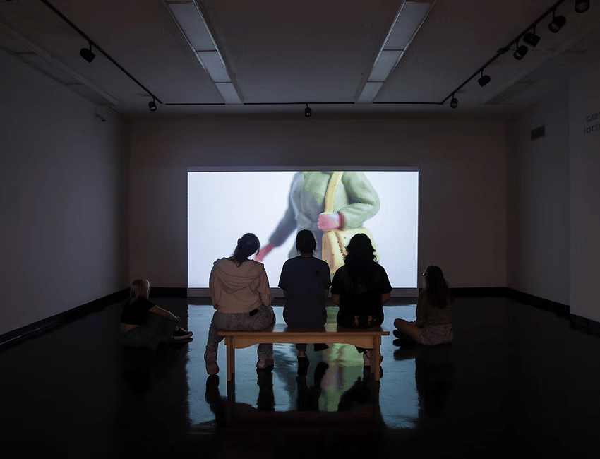 People watching a video installation. Video still of a plastic woman's torso, shown from waist to neck, carrying a handbag.