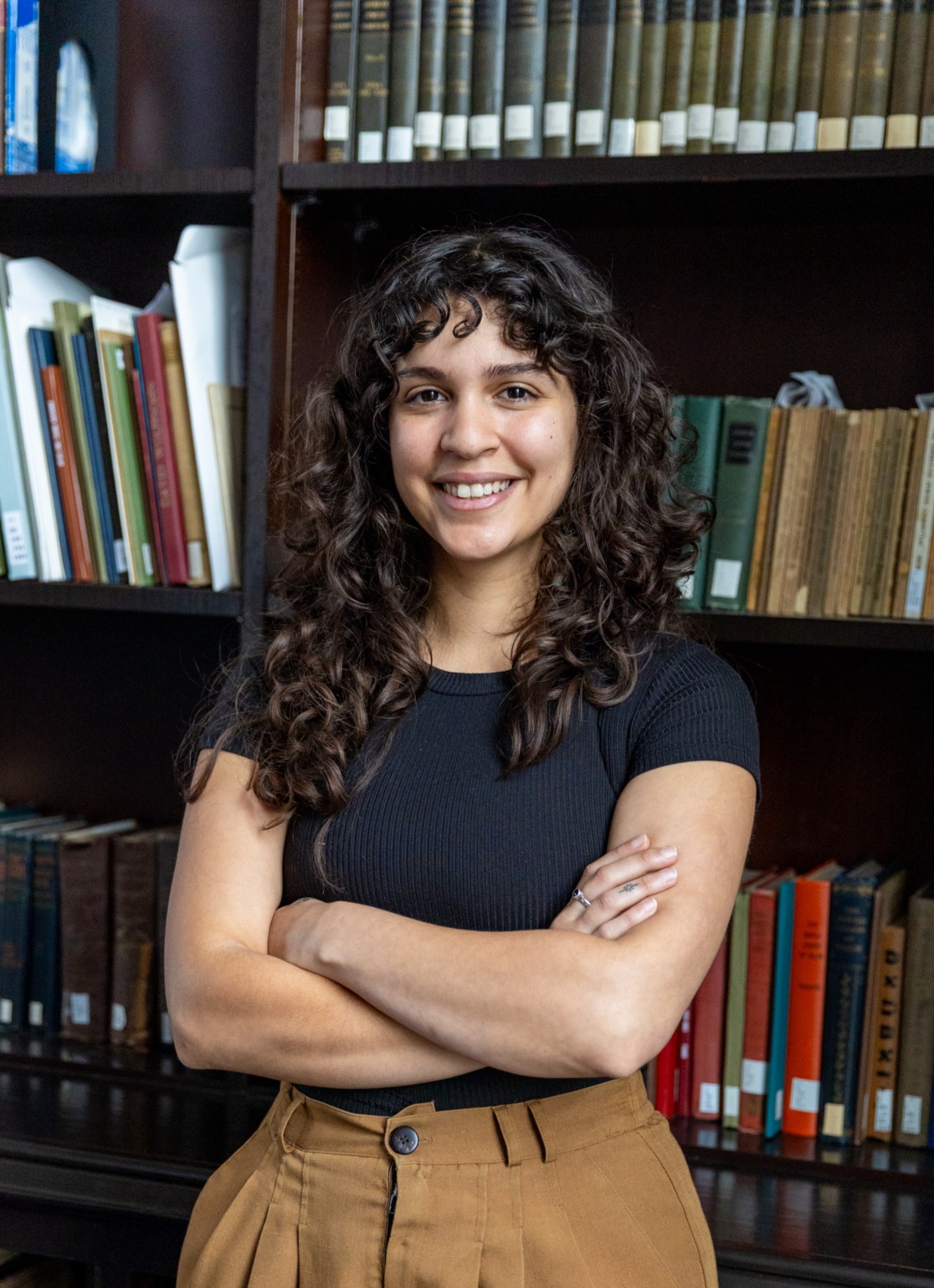 Headshot photo of Daniela Suzarte leaning against a library bookshelf.