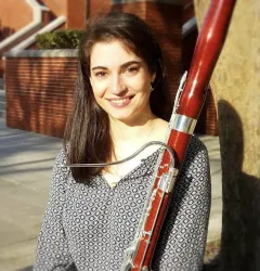 Image of Kayleigh holding a bassoon. Kayleigh has medium length brown hair and a patterned black and white blouse.