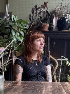 Image of Kodie. Kodie is staring off into the distance. Kodie is wearing a flannel tank top over a black shirt and Kodie has medium length red hair with bangs.