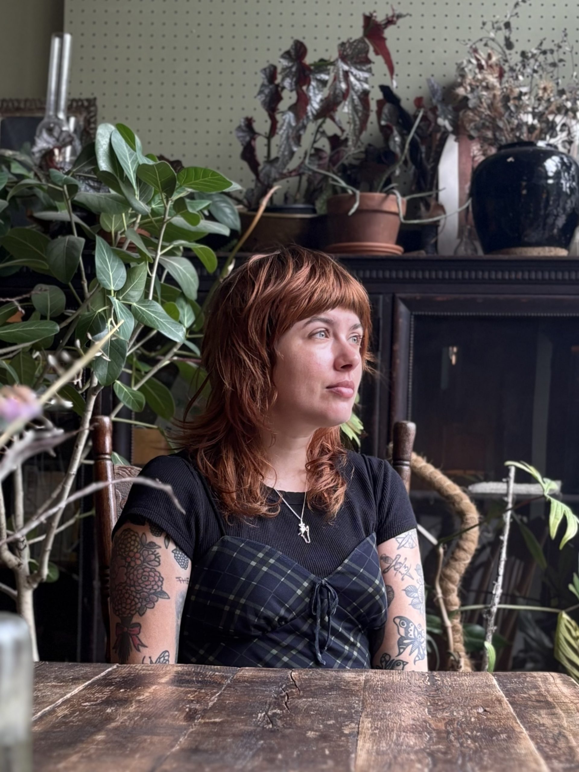 Image of Kodie. Kodie is staring off into the distance. Kodie is wearing a flannel tank top over a black shirt and Kodie has medium length red hair with bangs.