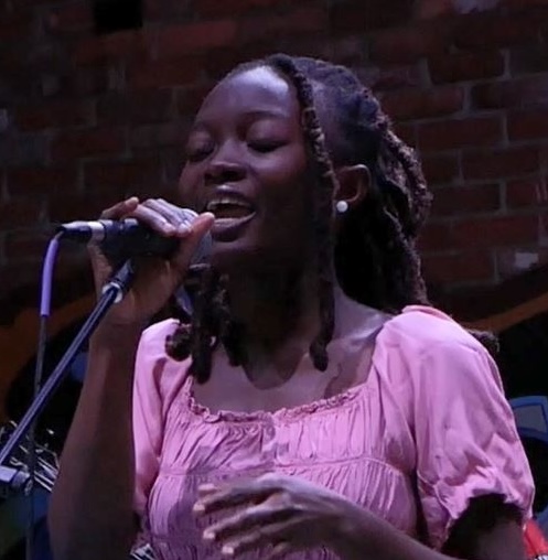 Image of Naa Akle while singing on stage. Naa Akle has shoulder length brown hair and a pink dress on