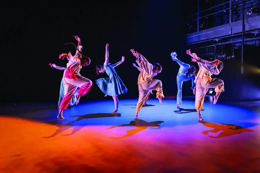 Dancers perform in a synchronous circle with arms outstretched and one leg lifted in forward motion