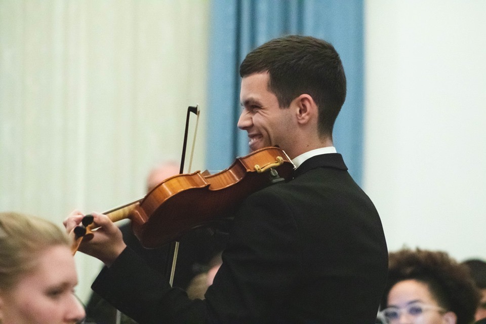 Side profile image of Brandon playing the violin. Brandon is wearing a dark suit and has short brown hair.