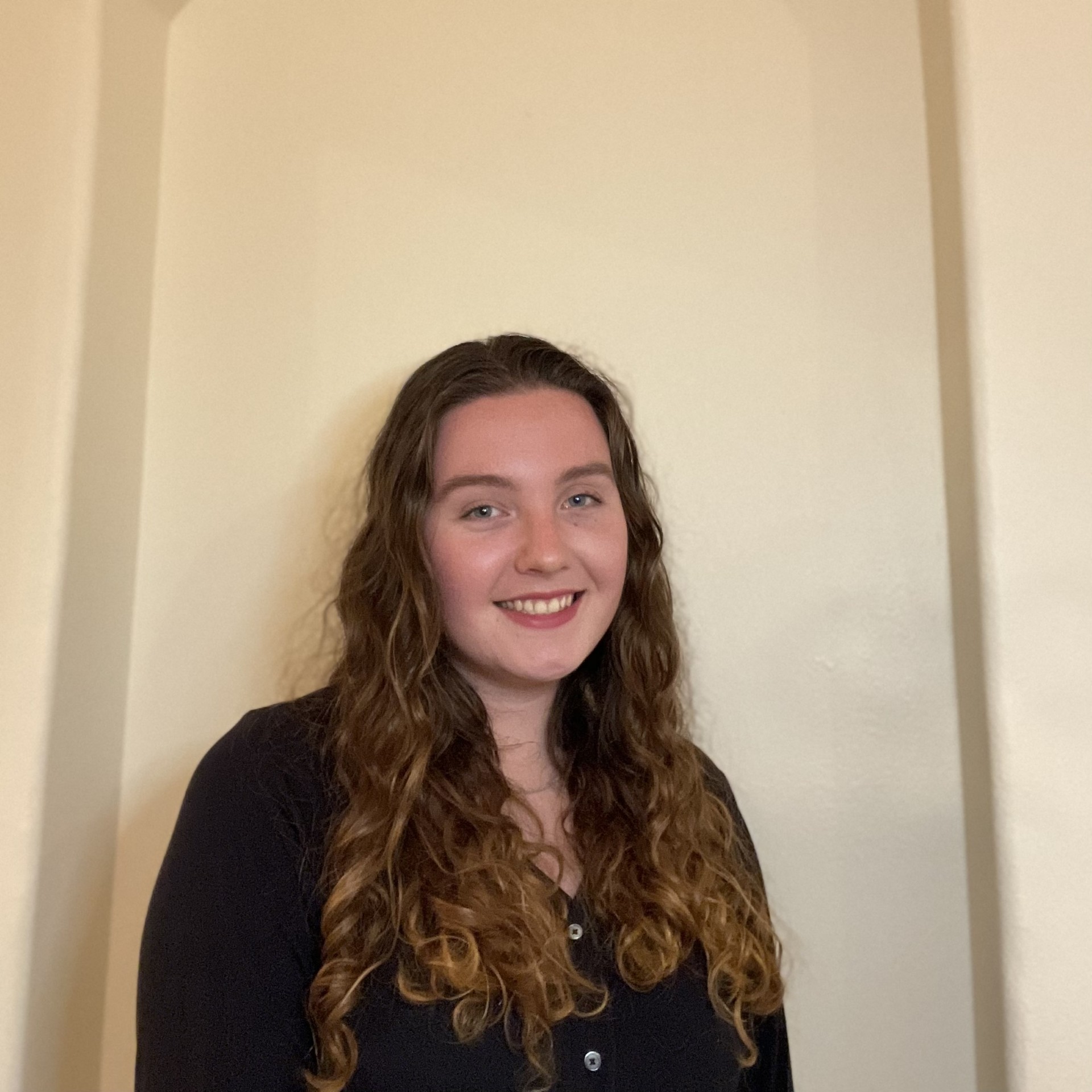 Image of Kaitlin. Kaitlin has long curly brown hair and is wearing a black shirt.