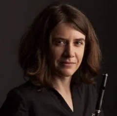 Image of Jeana holding a black flute with the top of the flute pointing up. She has shoulder length brown hair and a black shirt. She is looking right into the camera