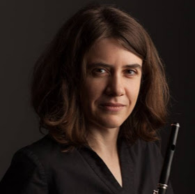Image of Jeana holding a black flute with the top of the flute pointing up. She has shoulder length brown hair and a black shirt. She is looking right into the camera