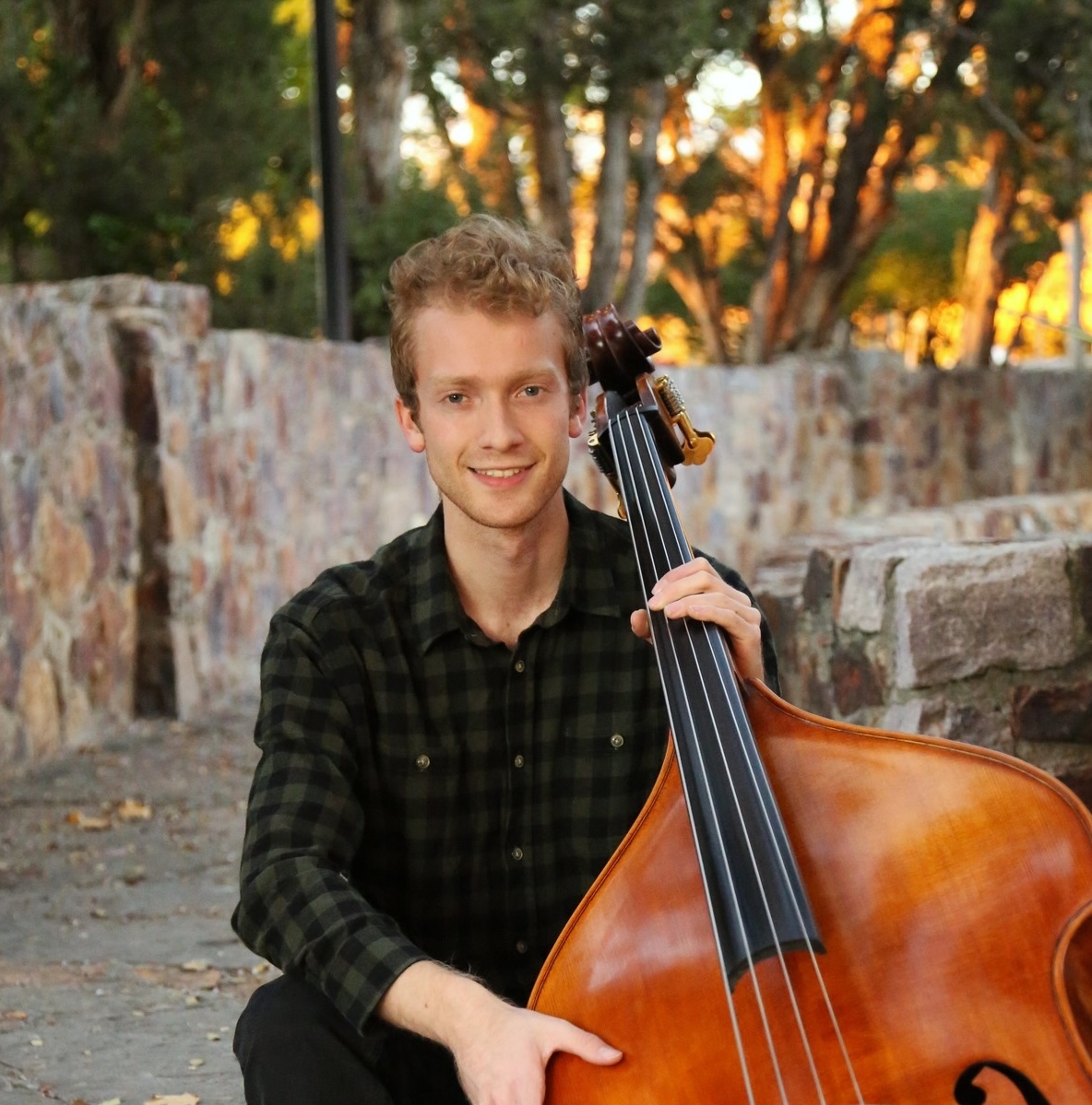 Image of Jans sitting, holding a stand up bass, wearing a green flannel and has short blonde hair, staring into the camera.