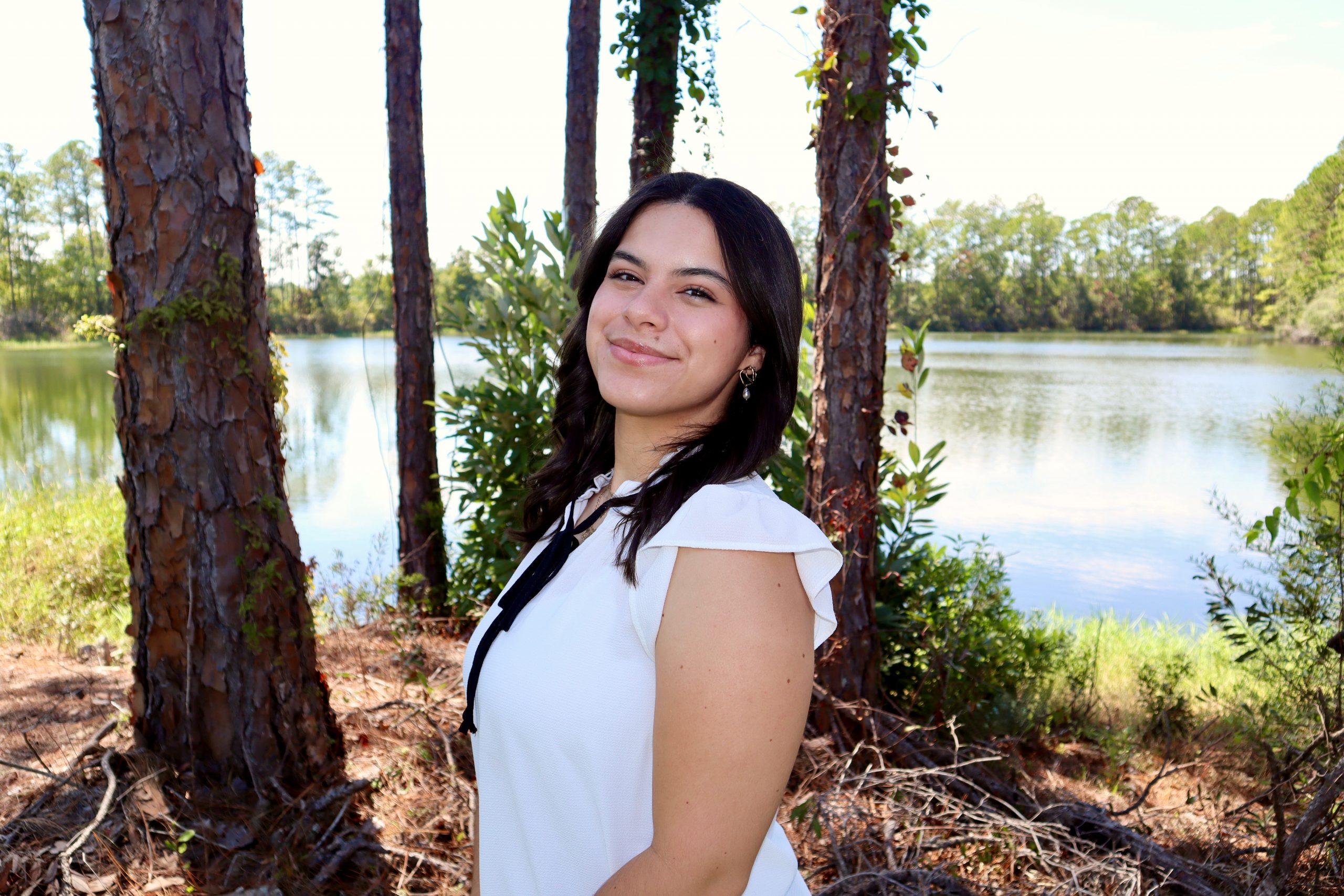 Karina Ocasio headshot photo, in an outdoor, lakeside setting with pine trees.