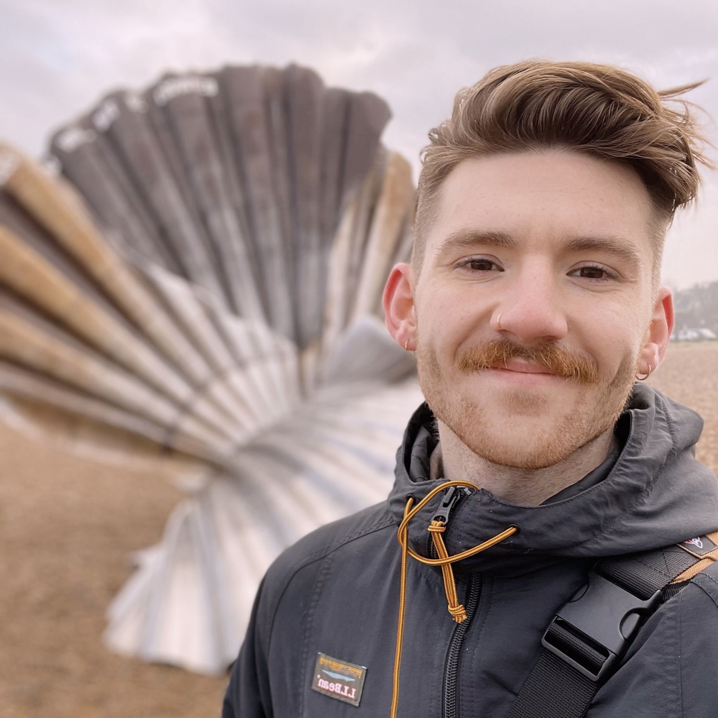 Selfie of Leo in front of a shell sculpture. Leo has short blonde hair and is wearing a dark grey jacket