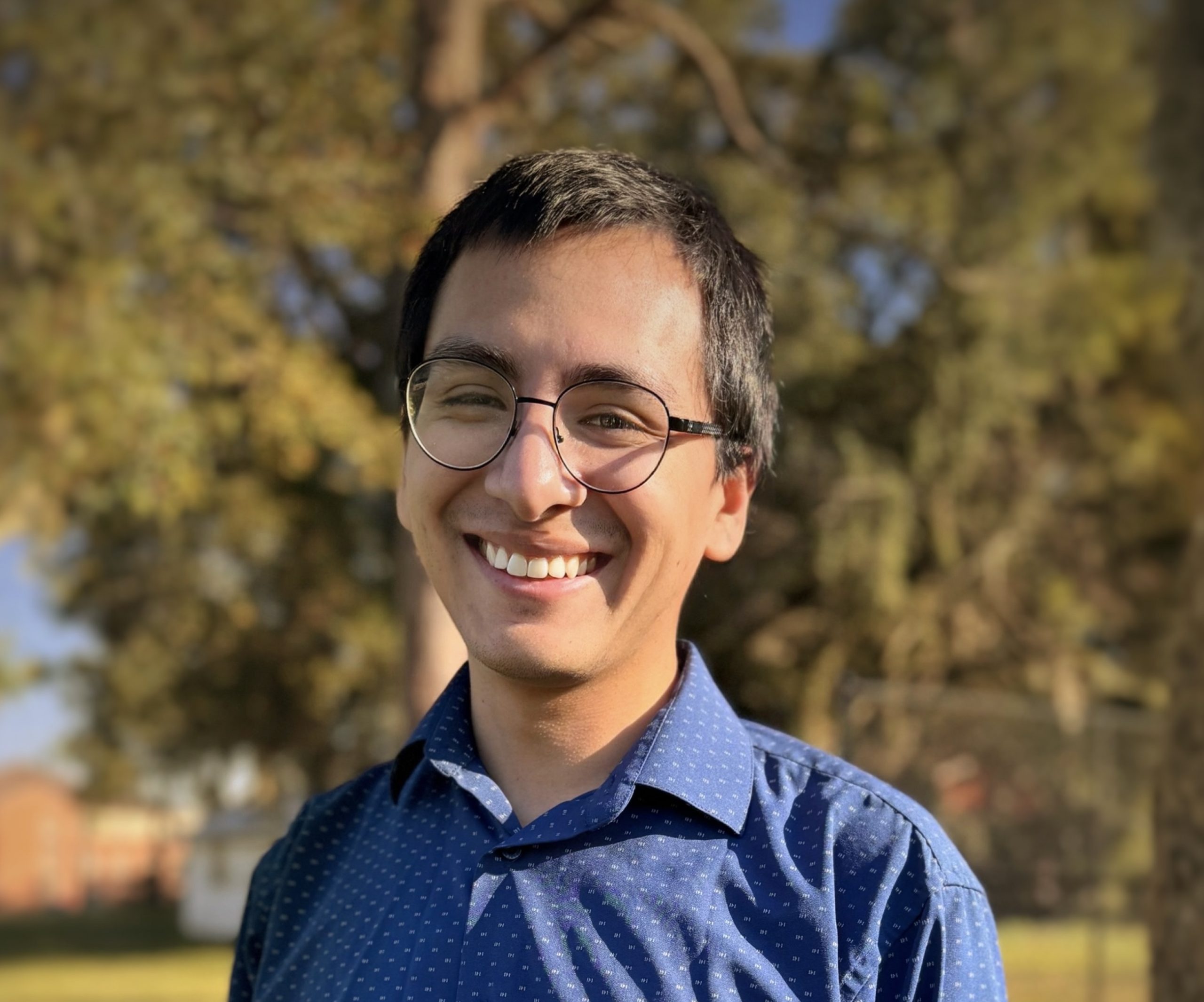 Image of Luis wearing metal rimmed glasses, a blue polo, and short black hair. Luis is looking into the camera