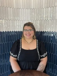 Image of Shelby. Shelby is wearing a black dress with white floral designs and has shoulder length brown hair