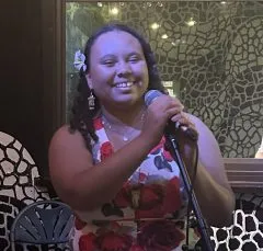 Image of Victoria in front of a microphone. Victoria is wearing a floral pattern dress and has medium length curly brown hair