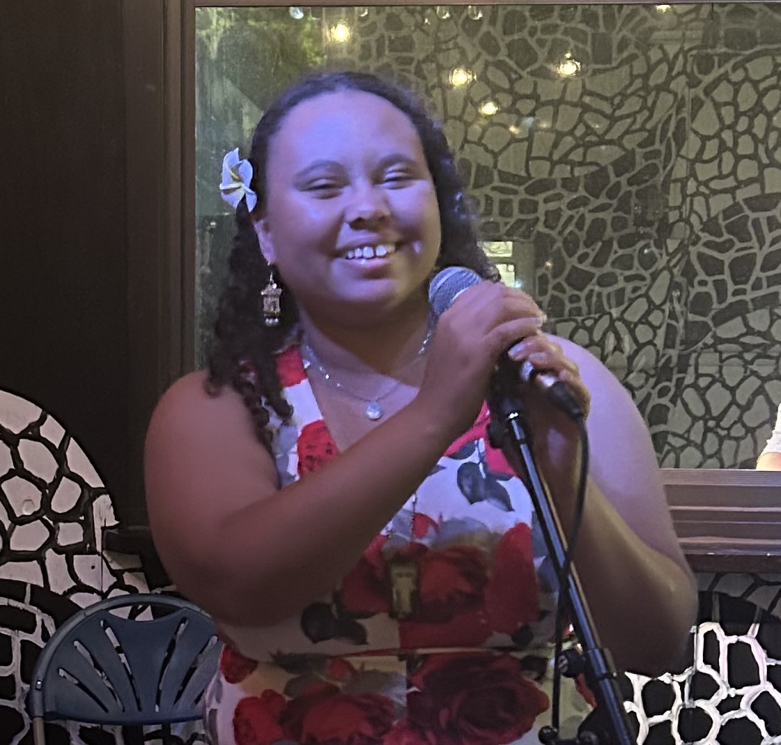 Image of Victoria in front of a microphone. Victoria is wearing a floral pattern dress and has medium length curly brown hair