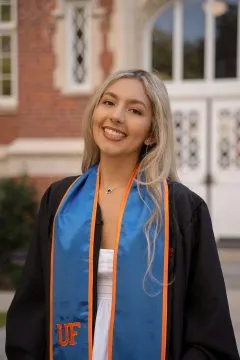 Image of Isabella. Isabella has long blonde hair and is wearing a UF graduation robe and stole over a white dress.