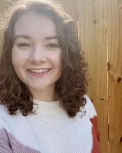 Selfie of Faith wearing a pink, orange, and white patterned sweater. Faith has curly shoulder length brown hair.